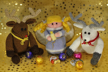 Gehäkelter Engel mit goldenen Flügeln zwischen einem braunen und einem hellen Amigurumi-Rentier vor goldenem Sternenhintergrund, davor verpackte Süßigkeiten.