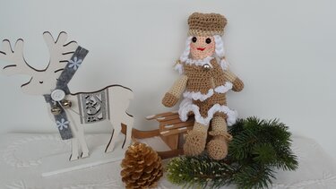 Gehäkelte Amigurumi-Puppe in Braun und Weiß auf Holzschlitten neben Holzrentier, Tannenzapfen und Tannenzweigen.