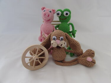 Gehäkeltes rosa Schwein, grüner Frosch und brauner Hase mit Wackelaugen auf einer kleinen Holzbank mit Holzrad.
