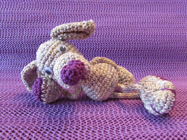 Gehäkelter Amigurumi-Hund in Creme mit lila Nase auf violettem, strukturiertem Stoff
