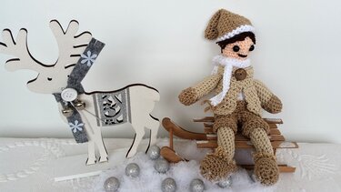 Beige-braune gehäkelte Amigurumi-Puppe mit Mütze und Schal auf Holzschlitten neben Holzrentier und silbernen Kugeln