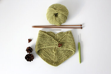 Materialien für Strickprojekt: grünes Garn, Stricknadeln, Häkelnadel, Stopfnadel und gestricktes Dreieckstuch