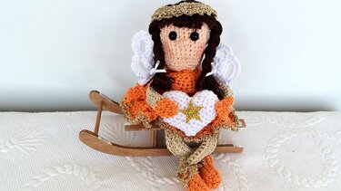 Gehäkeltes Engelchen-Amigurumi mit Flügeln sitzt auf einem Holzschlitten und hält ein Herz mit Stern