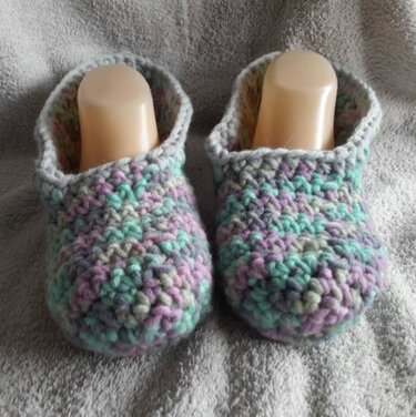 Gehäkelte Hausschuhe in Grau, Mint und Lila auf grauem, flauschigem Untergrund, mit Schuhformern.