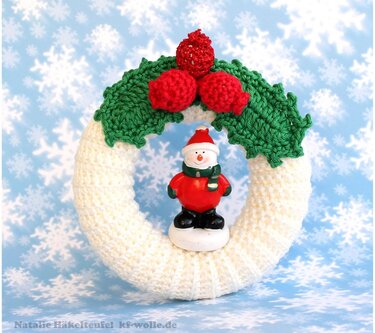 Gehäkelter Weihnachtskranz in Weiß und Grün mit Schneemann und roten Beeren vor Schneeflocken-Hintergrund.
