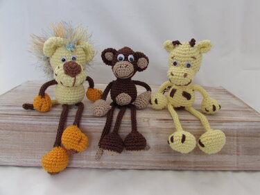 Drei gehäkelte Amigurumi-Tiere (Löwe, Affe, Giraffe) mit Wackelaugen auf Holz.