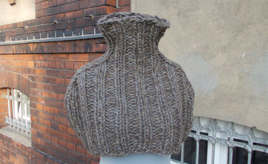 Grob gestrickter braun‑grauer Loop‑Schal mit Rippenmuster auf einer Kleiderbüste vor Backsteinwand.