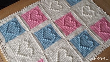 Crocheted baby blanket with pink, blue, and cream squares, each featuring a raised bobble-stitch heart.