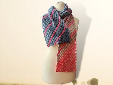 Gehäkelter Schal in Blaugrau, Beige und Koralle mit diagonalen pinken Streifen, an einer Schaufensterpuppe drapiert.