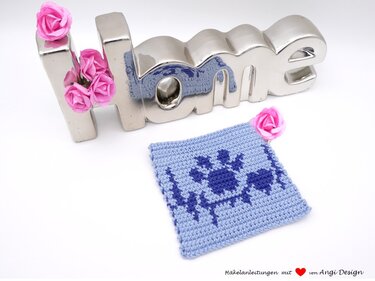 Quadratischer hellblauer geh
kelter Untersetzer mit dunkelblauem Pfotenabdruck- und Herzschlaglinien-Motiv, daneben silberner HOME-Schriftzug und rosa Deko-Blumen