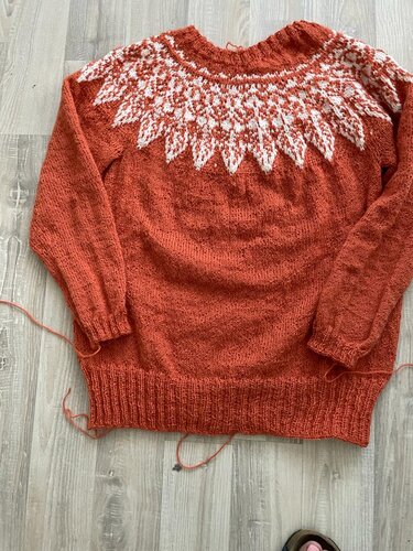 Rostroter Damenpullover mit weißer Fair-Isle-Rundpasse, flach liegend auf hellem Holzboden