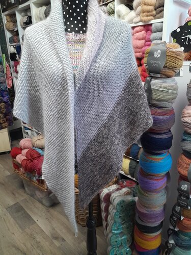 Gestricktes Schultertuch mit Grau-Farbverlauf auf einer Schaufensterpuppe, Garnregale im Hintergrund.