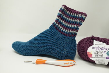 Handgehäkelte Socke in Petrol mit gestreiftem Bündchen, daneben Garnknäuel und Häkelnadel.