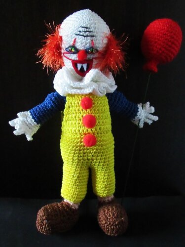 Gehäkelte Clownpuppe mit weißem Kopf, orangefarbenem Haar, gelber Latzhose und rotem Ballon vor schwarzem Hintergrund