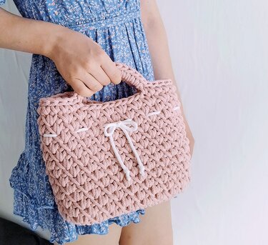 Rosafarbene, gehäkelte Henkeltasche mit weißem Kordelzug und strukturiertem Muster, gehalten von einer Person in einem blauen Kleid.