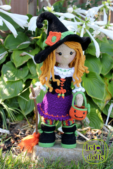 Crocheted amigurumi witch doll wearing black hat and purple skirt holding pumpkin bucket and broom, posed outdoors