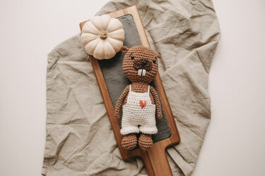 Gehäkeltes braunes Biber-Amigurumi mit weißer Latzhose und Herz, liegend auf Holzbrett neben kleinem weißen Kürbis