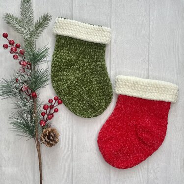 Zwei gehäkelte Weihnachtsstrümpfe, einer rot und einer grün, mit weißem Umschlag neben Tannenzweig, roten Beeren und Zapfen auf hellem Holz