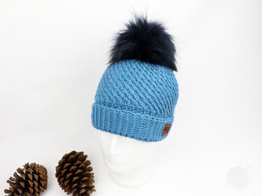Hellblaue gehäkelte Mütze mit dunklem Bommel und Rippenmuster auf weißem Kopfmodell, Tannenzapfen im Vordergrund