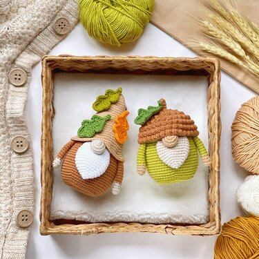 Two crocheted amigurumi autumn gnome dolls with leaf-decorated hats and white beards in a wicker tray, surrounded by yarn