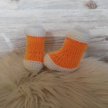 Gestrickte orange-weiße Babyschuhe auf weißem Kunstfell vor Holzwand.