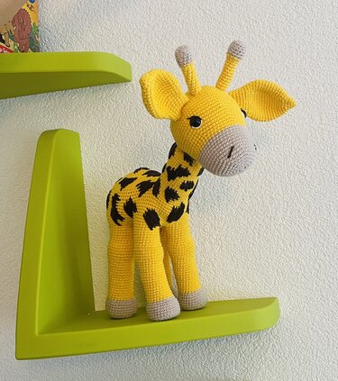Gehäkelte gelbe Amigurumi-Giraffe mit dunklen Flecken auf grünem Wandregal