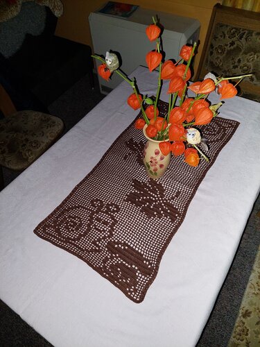 Dunkelbrauner gehäkelter Tischläufer mit Hirsch- und Waldmuster auf weißer Tischdecke, Vase mit orangen Lampionblumen und kleinen Eulenfiguren.