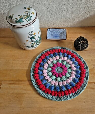 Runder mehrfarbiger Untersetzer mit Puff-Stichen auf Holztisch neben kleiner Schale