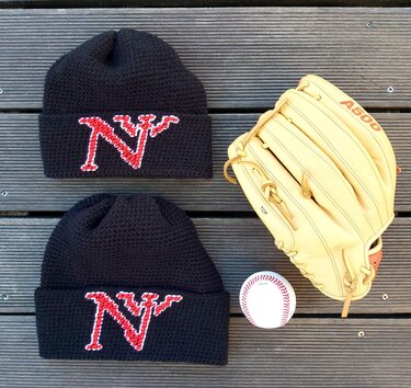Zwei handgehäkelte schwarze Beanies mit rotem N‑Motiv neben Baseballhandschuh und Baseball auf Holzlatten.