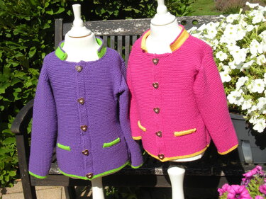 Two knitted children's cardigans on dress forms: purple with green trim and pink with yellow trim, shown outdoors on a bench.