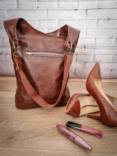 Braune Tasche mit außenliegendem Reißverschluss, Metallbeschlägen und Schulterriemen neben braunen Pumps und Make-up auf Holzboden.