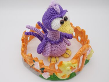 Gehäkelte lila und gelbe Amigurumi-Ente mit gelbem Schnabel, sitzend in einem orangefarbenen Deko-Zaun mit Kükenverzierung.