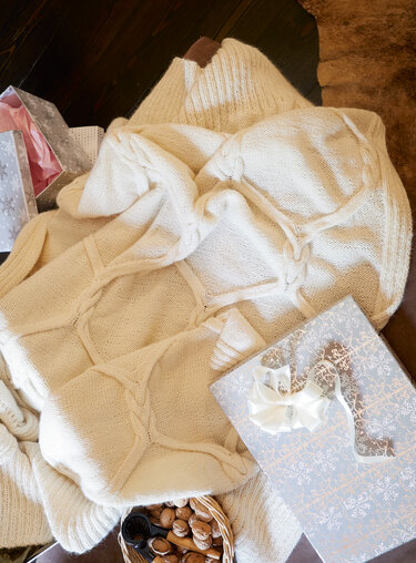Kuschelige cremeweiße Strickdecke mit Zopfmuster neben dekorativen Geschenkboxen und einem Korb mit Nüssen