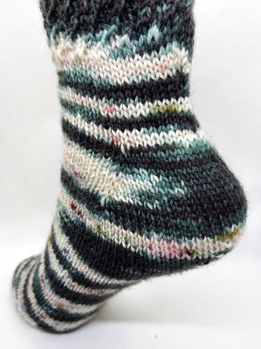 Nahaufnahme einer handgestrickten Socke mit dunkelgrauen, creme- und mintgrünen Streifen auf hellem Hintergrund.