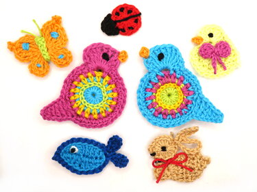Seven brightly colored crocheted appliques: pink bird, blue bird, orange butterfly, red ladybug, yellow chick with pink bow, blue fish, and tan bunny on a white background.