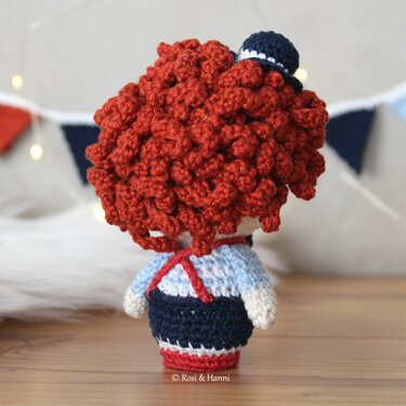 Rückseite einer Amigurumi-Puppe mit dichtem roten Lockenkopf, kleinem blauem Hut und gestreifter Kleidung vor unscharfer Girlande und Lichterkette.
