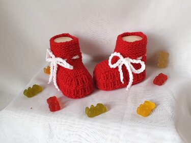 Zwei rote gestrickte Babyschuhe mit weißen Schleifenbändern auf hellem Stoff, dekoriert mit bunten Fruchtgummis