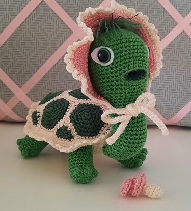 Gehäkelte grüne Amigurumi-Schildkröte mit rosa-weißem Häubchen und kleinem Schnuller