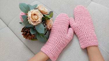 Rosa, grob gehäkelte Fäustlinge an den Händen neben Rosen und Tannenzapfen