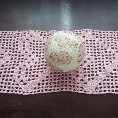 Rosafarbener filetgehäkelter Tischläufer mit wiederholten Herzmotiven und dekorativer Blumenkugel auf dunklem Untergrund