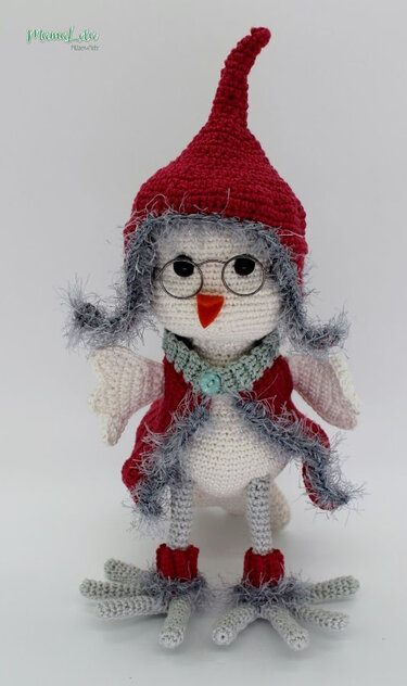 Gehäkelter Amigurumi-Vogel mit Brille, roter Zipfelmütze, Kragen, rotem Umhang und orangefarbenem Schnabel vor hellem Hintergrund
