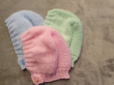 Drei handgestrickte Babymützen in Rosa, Hellblau und Mint mit kleinen Knöpfen auf grauem Stoff