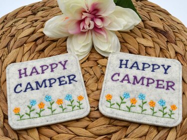 Zwei helle Filz-Tassenteppiche, angefertigt mit der ITH-Stickdatei, zeigen 'HAPPY CAMPER' in lila und blau sowie eine Reihe blauer und oranger Blumen auf einem geflochtenen Naturuntergrund.
