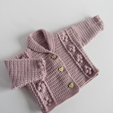 Dusty pink crocheted baby jacket with shawl collar and three heart-shaped buttons, flat-lay