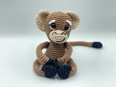 Small sitting amigurumi monkey in brown and beige yarn with large glossy eyes and a stitched smile; black hands, feet, and tail tip.