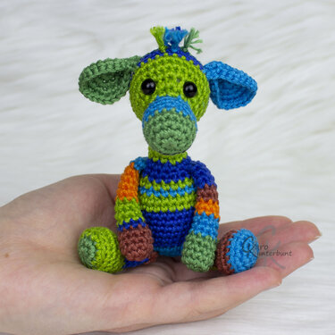 Hand hält kleines gehäkeltes Amigurumi in Blau-, Grün- und Orangetönen mit markanten Ohren