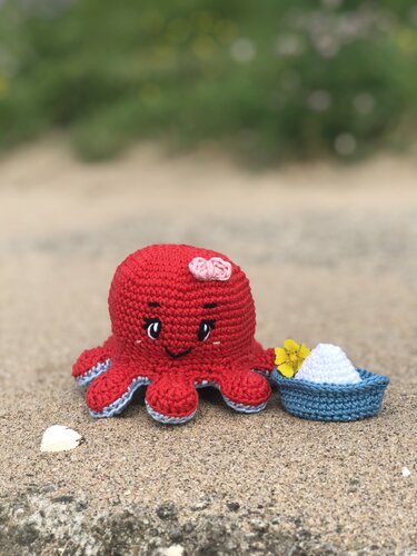 Gehäkelter roter Oktopus mit rosa Schleife, blauer Schale, gelber Blüte und Muschel auf sandigem Boden
