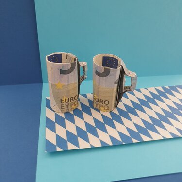 Zwei Origami-Bierkrüge aus Euro-Banknoten auf blau-weiß kariertem Untergrund