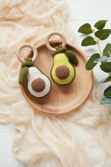 Two crocheted avocado baby rattles, one green and one cream, each attached to a wooden ring on a wooden tray with soft fabric and green leaves.