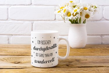 Weiße Keramiktasse mit Spruch „Ein bisschen durchgeknallt ist ganz nah an wundervoll“ neben Vase mit Gänseblümchen auf Holztisch
